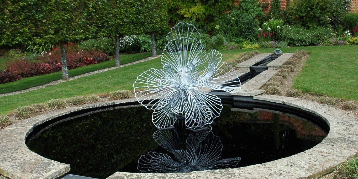 A large Flora sculpture installed in the middle of a circular garden pond making reflections in the water.