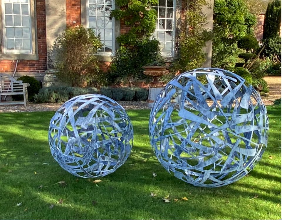 A 90cm and a 120cm diameter Calligraphy Ball sculpture in zinc coated steel on a lawn with a Georgian building behind.