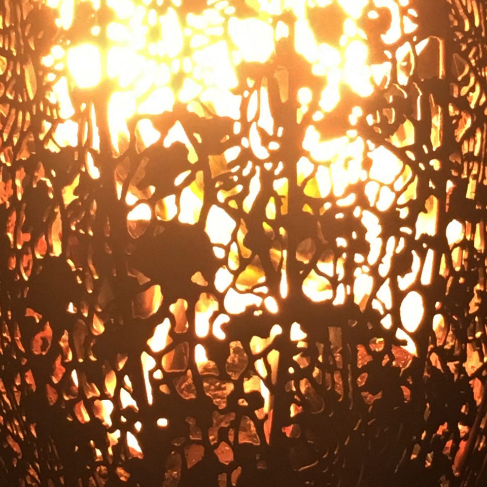 A detail of the metal patterns in the Filigree Fire Basket seen with fire alight behind the filigree metal wall.