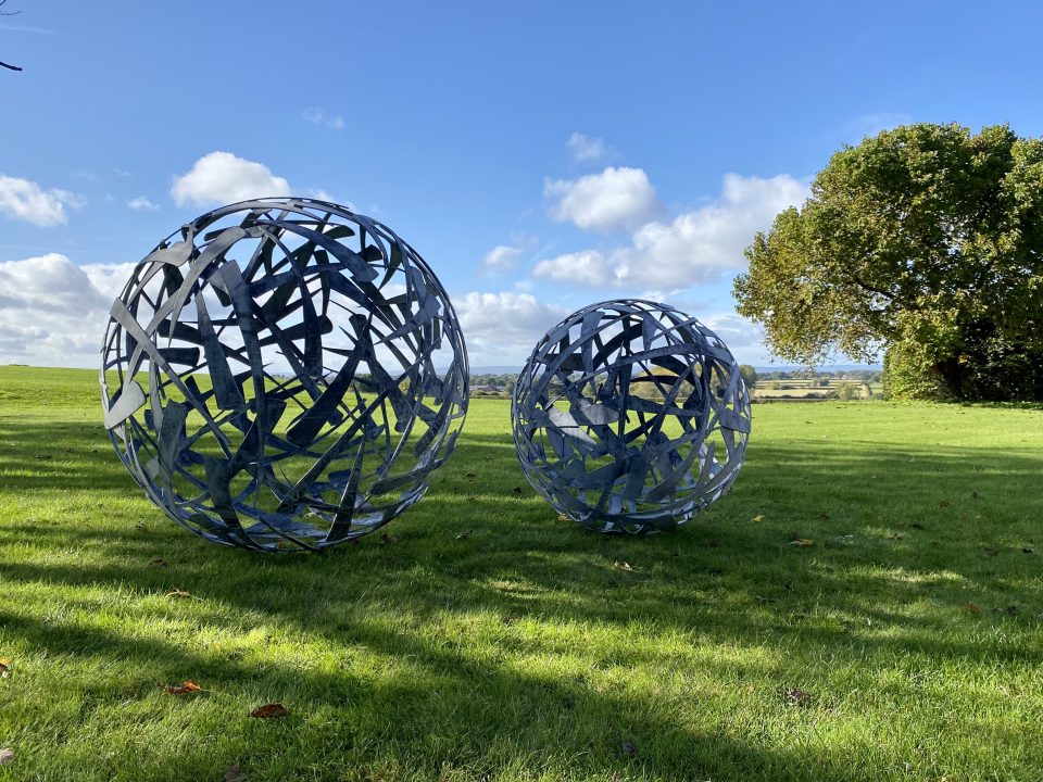 A 90cm and a 120cm diameter Calligraphy Ball sculpture in zinc coated steel on a lawn with a distant landscape view behind.