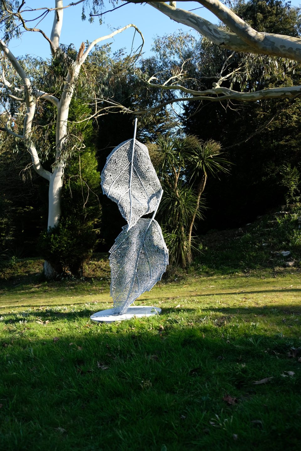 Falling Leaves sculpture in an open grassed area set on a base plate above ground seen againt a dark background of trees.