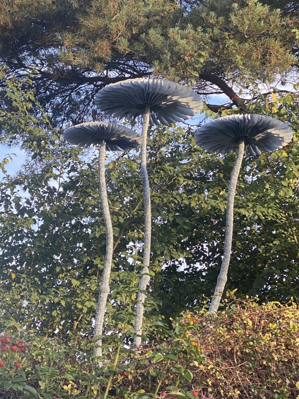 Detail of three metal mushroom sculptures seen from below against trees and sky.